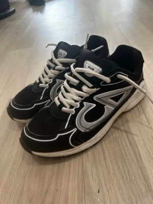 Svarta CD sneakers med vita detaljer - Snygga svarta sneakers från CD med vita och grå detaljer samt vita skosnören. Skorna har en sportig siluett med mesh och syntetmaterial, samt tydliga CD-loggor på sidorna. Perfekta för dig som vill sticka ut med exklusiva och trendiga sneakers.