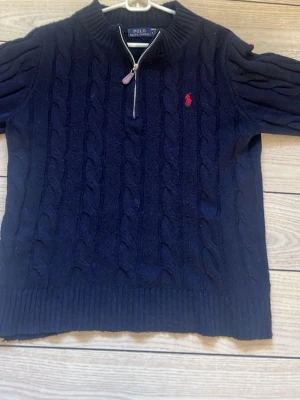 Mörkblå kabelstickad tröja Polo - Mörkblå kabelstickad tröja från Polo Ralph Lauren med halv dragkedja och röd broderad logga på bröstet. Nu vill jag helst bli av med den pg av att den har blivit för liten, inga problem med den och kan tvättas upp om någon vill