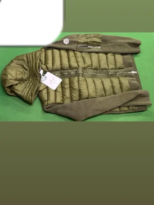 Olivgrön dunväst med huva Moncler - Snygg olivgrön dunväst från Moncler med stickade ärmar och huva. Västen har quiltad front, dragkedja och en liten ficka med dragkedja på ärmen. Perfekt för lager-på-lager och streetwear-stil.
