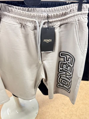 Beige shorts från Fendi - Snygga beige shorts från Fendi med elastisk midja och snörning. Stora Fendi-loggan tryckt längs ena benet ger en cool vibe. Perfekta för sommaren och chill dagar. Mjukt material och praktiska fickor på sidorna.