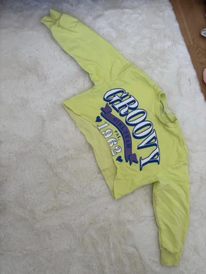 Gul cropped hoodie med tryck - Cool gul cropped hoodie med stort blått och lila tryck på framsidan där det står 'GROOVY GIRL CLUB 1982' och små hjärtan. Hoodien har långa ärmar och ribbade muddar. Perfekt för dig som gillar färgglada statement-plagg.