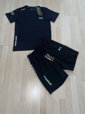 Under Armour träningsset marinblå sett  - Snyggt träningsset från Under Armour med t-shirt och shorts. Finns i svart och blått, båda med färgglada logotyper och text på sidan. Materialet är lätt och andas, perfekt för gym eller löpning. T-shirten har rund hals och korta ärmar, shortsen har elastisk midja.