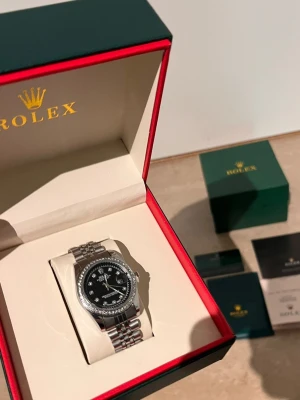 Role Datejust silver klocka - Snygg Role Datejust med silverfärgat armband i metall och svart urtavla. Klockan har en bezel med glittrande stenar och klassisk jubilee-länk. Tydlig datumvisning och eleganta indexmarkeringar. Perfekt accessoar för dig som vill sticka ut.