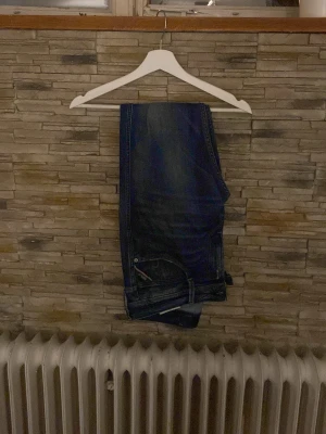 Mörkblå jeans med klassisk passform - Säljer ett par mörkblå jeans med klassisk passform och snygga detaljer vid fickorna. Jeansen har raka ben och är tillverkade i denim. Perfekta för dig som gillar en tidlös stil och vill ha ett par jeans som funkar till det mesta.