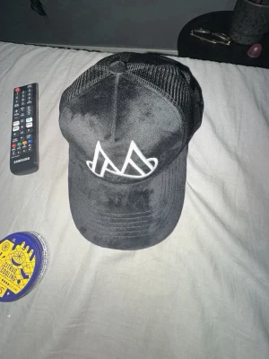Svart meshkeps med vit logga - Säljer en svart keps med mesh på sidorna och bak, samt en böjd skärm. Framtill finns en stor vit broderad logga i form av två stiliserade M. Kepsen har justerbar snapback och är tillverkad i en blandning av tyg och mesh för skön ventilation.