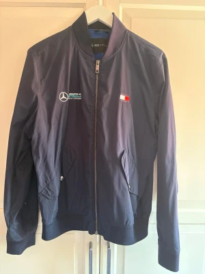 Mörkblå Mercedes Tommy Hilfiger bomber - Snygg mörkblå bomberjacka från Tommy Hilfiger med Mercedes AMG Petronas-logga på bröstet. Jackan har dragkedja, två fickor med tryckknapp och ribbade muddar. Materialet är tunt och lätt, perfekt för vår och höst.