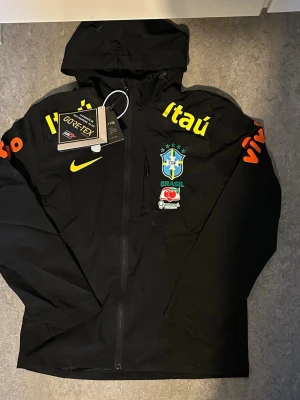 Nike Brasil svart vindjacka med huva - Svart vindjacka från Nike med Brasilien CBF-logga, gula och orangea sponsortryck samt Guaraná Antarctica-märke. Jackan har huva, hel dragkedja och är tillverkad i GORE-TEX-material som skyddar mot vind och regn. Perfekt för dig som gillar sportig stil och fotboll.