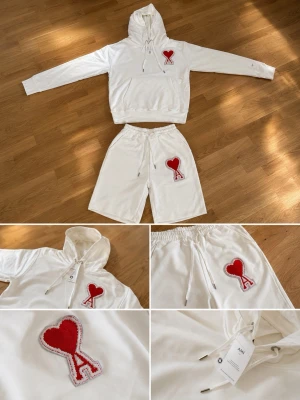 Vit hoodie från AMI med rött hjärta - Säljer en clean vit hoodie från AMI med stor röd hjärtlogga och bokstaven A på bröstet. Hoodien har huva med dragsko, känguruficka och ribbade muddar. Tillverkad i mjuk bomull för skön känsla. Perfekt för dig som gillar streetwear och vill sticka ut.