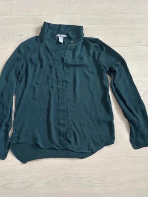 Mörkgrön skjorta från Filippa K - Stilren mörkgrön skjorta från Filippa K med klassisk krage och knappar framtill. Skjortan har lång ärm och är tillverkad i ett mjukt, lätt material som ger en snygg och avslappnad look. Perfekt för dig som gillar minimalistisk stil.