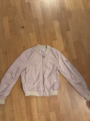 Ljusrosa bomberjacka med dragkedja - Snygg ljusrosa bomberjacka med ribbade muddar i beige, silverfärgad dragkedja framtill och fickor med tryckknappar. Ärmen har en extra ficka med dragkedja för cool detalj. Jackan är i ett glansigt material som ger en modern vibe.