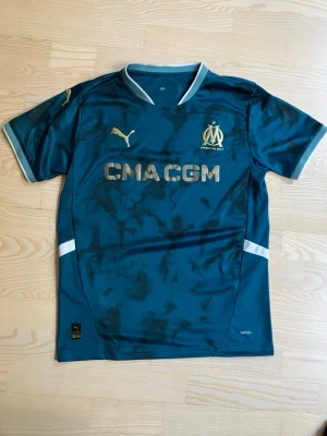 Marseille matchtröja Puma blå/guld - Snygg Marseille matchtröja från Puma i mörkblått med gulddetaljer och klubbmärke på bröstet. Tröjan har korta ärmar, V-ringning och tryckt sponsorlogga framtill. Tillverkad i lätt och ventilerande material, perfekt för fotbollsträning.