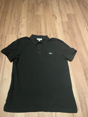 Svart pikétröja från Lacoste - Klassisk svart pikétröja från Lacoste med korta ärmar och krage. Tröjan har den ikoniska gröna krokodilloggan broderad på bröstet och knäppning framtill. Perfekt till jeans eller shorts för en clean och stilren look.