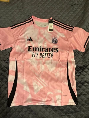Rosa Real Madrid tröja från Adidas - Snygg rosa Real Madrid matchtröja från Adidas med svart logga och klubbmärke på bröstet. Tröjan har svarta detaljer, tre ränder på axlarna och texten 'Emirates Fly Better' på framsidan. Tillverkad i lätt och ventilerande material med AEROREADY-teknologi.