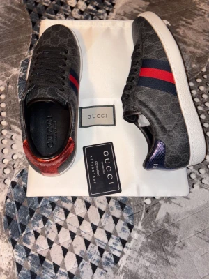 Gucci Ace sneakers med GG-mönster - Gucci Ace sneakers i svartgrått GG-mönstrat tyg med klassiska röda och blå ränder på sidan. Skorna har vit sula och metallic-detaljer i rött och blått på hälen med Gucci-logga. Snygg och ikonisk design som sticker ut.