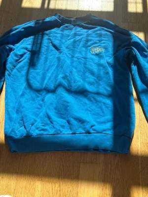 Blå sweatshirt från junkyard  - En blå sweatshirt från junkyard med rund halsringning och ribbade muddar vid ärmslut och nederkant. Tröjan har en liten broderad logga på bröstet och är långärmad. Perfekt för en avslappnad och stilren look.