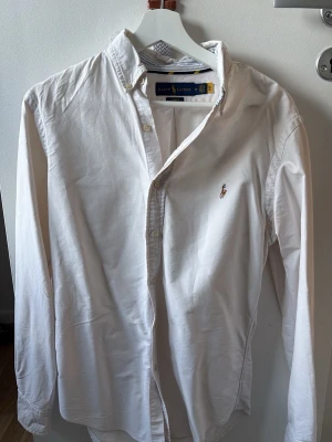 Vit slim fit skjorta från Ralph Lauren - Vit långärmad skjorta från Ralph Lauren i slim fit-modell. Skjortan har klassisk krage, knäppning framtill och den ikoniska broderade logotypen på bröstet. Materialet är bomull och designen är stilren och tidlös.