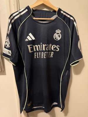 Real Madrid away jersey 2025/2026 authentic  - Mörkblå Real Madrid bortatröja från Adidas med vita detaljer och klubbmärke på bröstet. Tröjan har tryckta sponsorer och Champions League-märke på ärmen. Tillverkad i lätt och ventilerande funktionsmaterial, perfekt för fotbollsträning eller match.