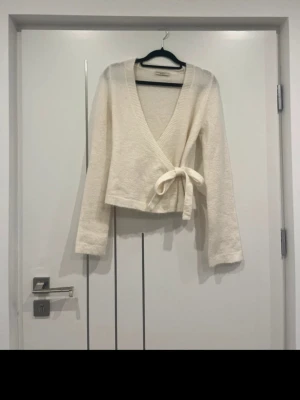 Vit omlottstickad tröja - Säljer en krämvit stickad omlottopp med långa ärmar och knytband i midjan. Tröjan har djup V-ringning och ribbstickade kanter. Perfekt för lager på lager och enkel att styla till olika outfits.