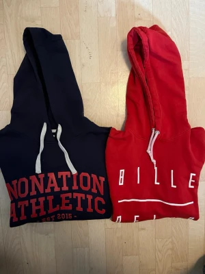 Två hoodies i rött och mörkblått - Säljer två hoodies, en mörkblå från Nonation Athletic med röd text och vita snören, samt en röd hoodie från Bille med vit text och snören. Båda har huva och klassisk passform, perfekta för en avslappnad look.