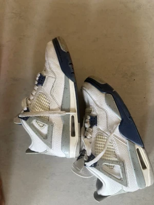 Air Jordan 4 Retro White Navy - Snygga Air Jordan 4 Retro sneakers i vitt läder med detaljer i navyblått och ljusgrått. Klassisk mesh på sidorna och tunga, samt ikonisk Jumpman-logga på plösen. Skorna har en robust och sportig design med högt skaft och synlig Air-dämpning i sulan.