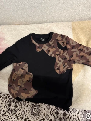 Fendi brun och svart sweatshirt - Cool svart sweatshirt från Fendi med bruna och beige camouflageinspirerade partier och FF-logga över hela mönstret. Tröjan har rund halsringning och långa ärmar. Perfekt för dig som vill sticka ut med designerstil.