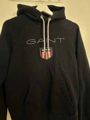 Mörkblå hoodie från GANT med logga - Snygg mörkblå hoodie från GANT med broderad logga och amerikanskinspirerat sköldmärke på bröstet. Hoodien har vit dragsko i huvan och en stor magficka framtill. Perfekt för en avslappnad och stilren look.