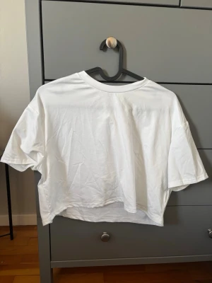 Vit croppad t-shirt i bomull - En basic vit croppad t-shirt med rund hals och korta ärmar. Oversized passform som ger en avslappnad look. Tillverkad i mjuk bomull, perfekt för en enkel och stilren outfit.