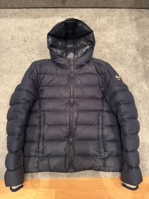 Colmar dunjacka - Snygg mörkblå dunjacka från Colmar Originals med huva och quiltad design. Jackan har dragkedja framtill och en diskret logga på ärmen. Perfekt för kalla vinterdagar och riktigt skön att bära. Klassisk modell som funkar till det mesta.