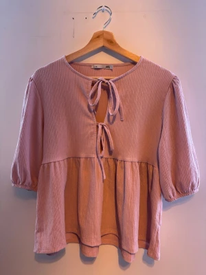 Ljusrosa blus med knyt  - Supersöt ljusrosa blus från lager 157 med tre knytband framtill och lätt puffade trekvartsärmar. 