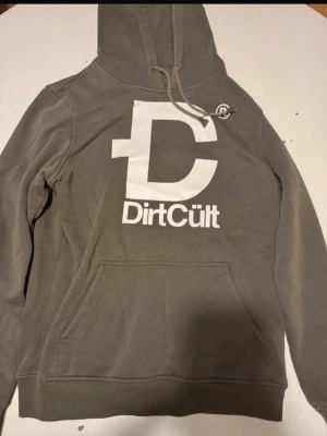 Olivgrön DirtCült hoodie med tryck - Säljer en olivgrön hoodie från DirtCült med stor vit logga och text på bröstet. Hoodien har huva med dragsko och en stor magficka. Tillverkad i mjukt bomullsmaterial, perfekt för chill dagar. Snygg streetwear vibe och enkel att matcha.