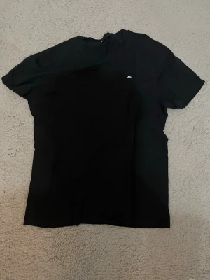 Svart t-shirt från J.Lindeberg - En stilren svart t-shirt från J.Lindeberg med liten vit logga på bröstet. Klassisk passform och korta ärmar, perfekt till jeans eller shorts. Gjord i mjuk bomull för en skön känsla hela dagen.