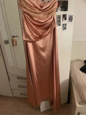 Lång glittrig rosa balklänning - Elegant långklänning i skimrande rosa satin med draperad överdel och glittriga detaljer vid axlarna. Klänningen har en vacker omlottkänsla upptill och faller mjukt längs kroppen. Perfekt för bal eller festliga tillfällen.