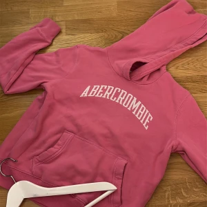 Rosa hoodie från Abercrombie Kids - Säljer en rosa hoodie från Abercrombie Kids med stor vit logga på bröstet. Hoodien har en klassisk känguruficka framtill och mjuk insida i fleece. Perfekt för dig som gillar en avslappnad och bekväm stil. Storlek 11/12 men passar xxs