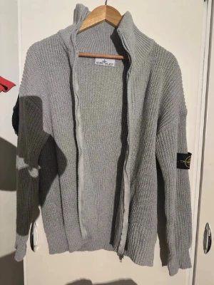 Grå stickad ziptröja Stone Island - Säljer en grå stickad ziptröja från Stone Island med hög krage och ribbstickad struktur. Tröjan har dragkedja framtill och den klassiska Stone Island-loggan på vänster ärm. Perfekt för dig som gillar streetwear och snygga detaljer. Kom privat för mer bilder!