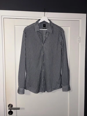 Randig skjorta från Hugo Boss - Snygg långärmad skjorta från Hugo Boss med smala vertikala ränder i vitt och mörkblått. Skjortan har klassisk krage och knappar framtill. Regular fit-modell som passar perfekt till både jeans och chinos.