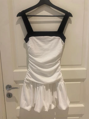 Vit kort klänning med svarta band - Säljer en vit kort klänning med breda svarta axelband och fyrkantig urringning. Klänningen har rynkade detaljer i midjan och en volangig kjol. Perfekt för dig som vill ha en stilren och trendig look. Använt ett få tal gånger💕