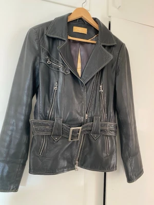 Svart skinnjacka med bälte  - Snygg svart skinnjacka från BIBA med bred krage, flera dragkedjor och ett justerbart och avtagbart bälte i midjan. Jackan har en klassisk bikerstil med detaljerade sömmar och silverfärgade metalldetaljer. Perfekt för dig som vill ha en cool och tidlös look.