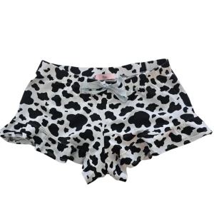 Svartvita shorts med ko-mönster och rosett - Superfina shorts med svartvitt ko-mönster och en söt ljusgrå rosett framtill. De har volangkant nedtill och elastisk midja för extra komfort. Perfekta för dig som gillar unika och lekfulla plagg.