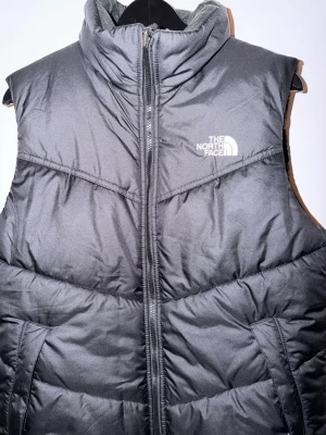 Svart dunväst från The North Face - Snygg svart dunväst från The North Face med hög krage och dragkedja framtill. Västen har två fickor på sidorna och logga på både bröstet och ryggen. Perfekt för lager-på-lager och håller dig varm under kyliga dagar. Lite bredare form