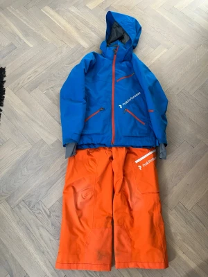 Orange täckbyxor från Peak Performance - Säljer ett par orange täckbyxor från Peak Performance med logga på vänster ben. Byxorna har bred passform och är tillverkade i slitstarkt, vattenavvisande material. Perfekta för skidåkning eller vinteraktiviteter.