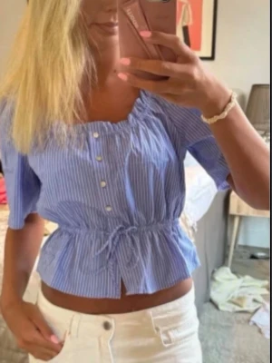 Blårandig blus från H&M - Säljer en söt blå- och vitrandig croppad blus med korta puffärmar, knappar framtill och knyt i midjan. Blusen har en elastisk, rynkad halsringning och volangkant nedtill. Perfekt för sommarens alla tillfällen!