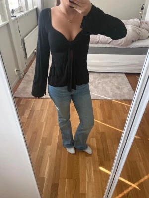 Svart långärmad topp från Design by si - Säljer en svart långärmad topp från Design by si med djup v-ringning och knyt framtill. Toppen har en figurnära passform och är tillverkad i mjukt material som känns skönt mot huden. Perfekt att styla med jeans eller kjol.