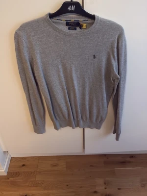 Grå stickad tröja från Polo Ralph Lauren - Klassisk grå stickad tröja från Polo Ralph Lauren med rund halsringning och diskret broderad logga på bröstet. Tröjan har långa ärmar och ribbade muddar vid ärmslut och nederkant. Perfekt basplagg i mjukt material.