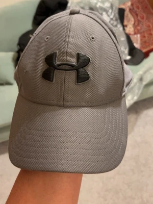 Grå keps från Under Armour - Snygg grå keps från Under Armour med svart broderad logga framtill och text bak. Kepsen har böjd skärm och är tillverkad i ett ventilerande syntetmaterial som passar perfekt till sport och vardag.
