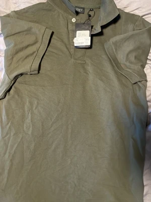 Olivgrön pikétröja från Lagency - Snygg olivgrön pikétröja från Lagency med klassisk krage och två knappar vid halsen. Tillverkad i mjuk bomull och har korta ärmar. Perfekt för en avslappnad och stilren look.