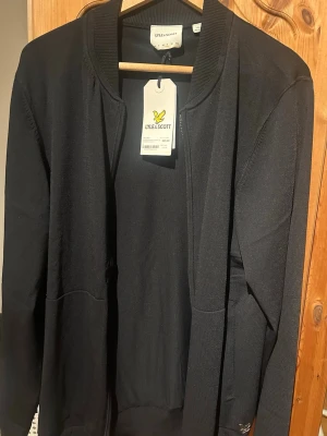 Svart ziptröja från Lyle & Scott - Snygg svart ziptröja från Lyle & Scott med ribbade muddar och ståkrage. Tröjan har dragkedja framtill och två fickor. Perfekt till lager på lager och enkel att matcha med olika outfits.