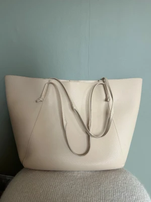 Stor beige shoppingväska i skinnimitation - Minimalistisk shoppingväska i beige skinnimitation med två långa axelband och enkel design utan synliga loggor. Väskan har en rymlig insida och mjukt, lätt strukturerat material. Perfekt för dig som gillar stilrena accessoarer och vill ha plats för allt du behöver.