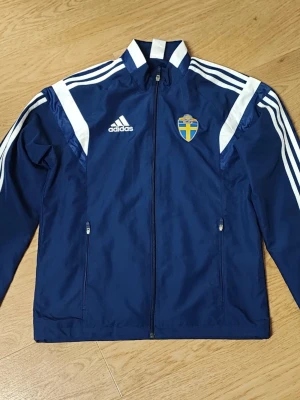 Blå Adidas Svff träningsset - Säljer ett blått träningsset från Adidas med Sveriges landslagsemblem på jackan. Setet består av en jacka med dragkedja och matchande byxor med vita ränder längs ärmar och ben. Byxorna har också det lillasye hålet på vänster knä