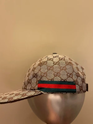  beige keps med GG-mönster - Säljer en beige keps  med klassiskt GG-monogram över hela kepsen. På sidan finns en grön och röd rand som ger den en ikonisk touch. Kepsen har böjd skärm och justerbar rem bak. Materialet är canvasliknande och ger en lyxig känsla.