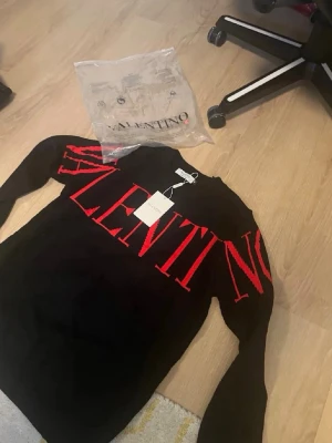 Svart stickad tröja från Valentino - Snygg svart stickad tröja från Valentino med stort rött VALENTINO-tryck över bröstet och armarna. Tröjan har rund halsringning och långa ärmar. Perfekt för dig som vill ha en statement piece i garderoben.